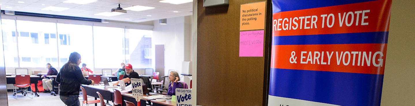 Photo of a room with people seated at long tables covered with forms and Vote Here signs. To the right is a banner with a blue background and white text on red stripes that reads Register to vote and early voting.