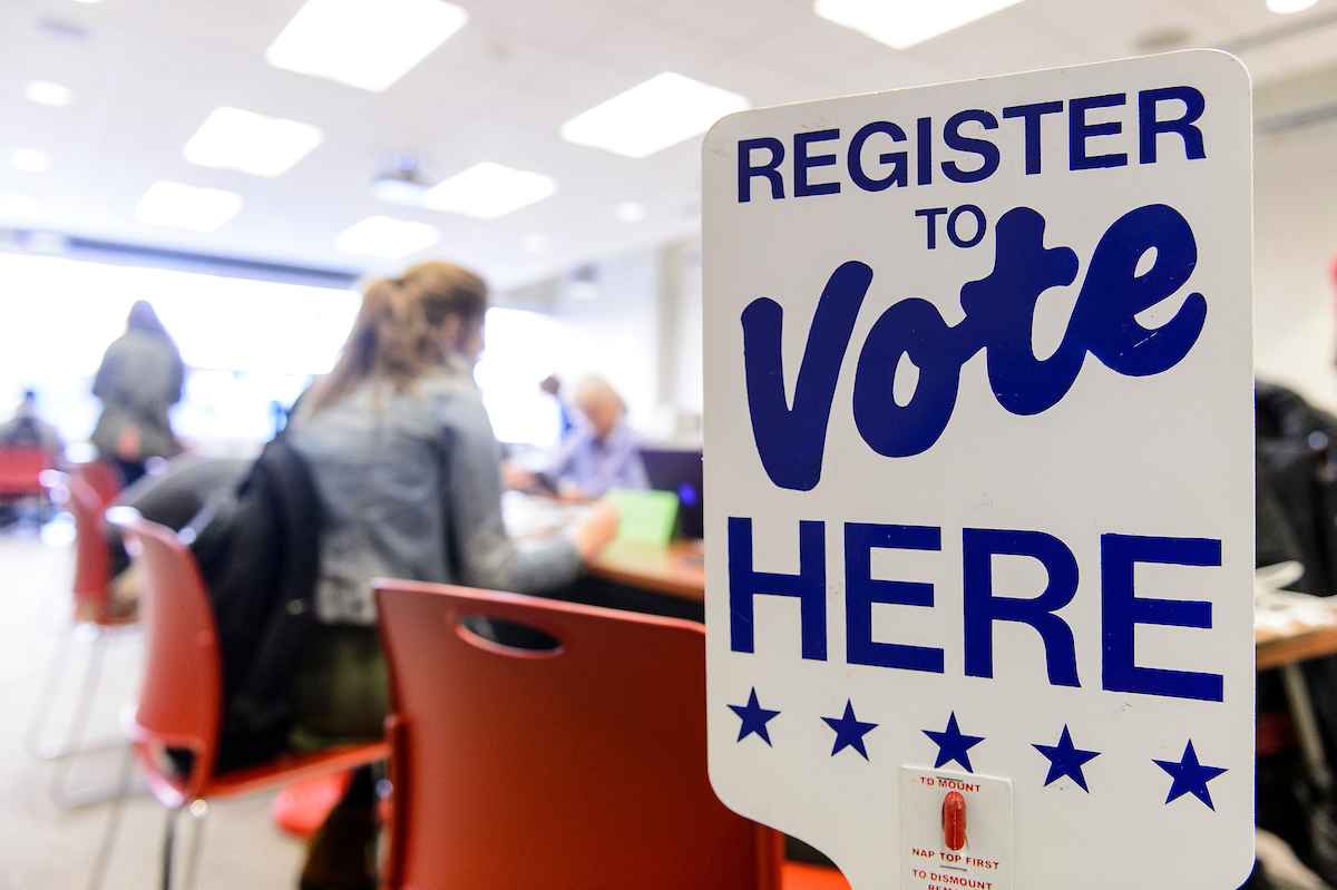Photo of a polling place. In the foreground, a white sign with blue text reads Register to vote here. In the background, people sit at tables with voter registration paperwork.
