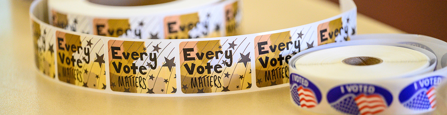 Photo of two rolls of stickers sitting on a table. The larger roll in the background has gold stickers with black stars and text that reads Every vote matters. The smaller roll in the foreground has blue oval stickers with an illustration of a U.S. flag and the words I voted.