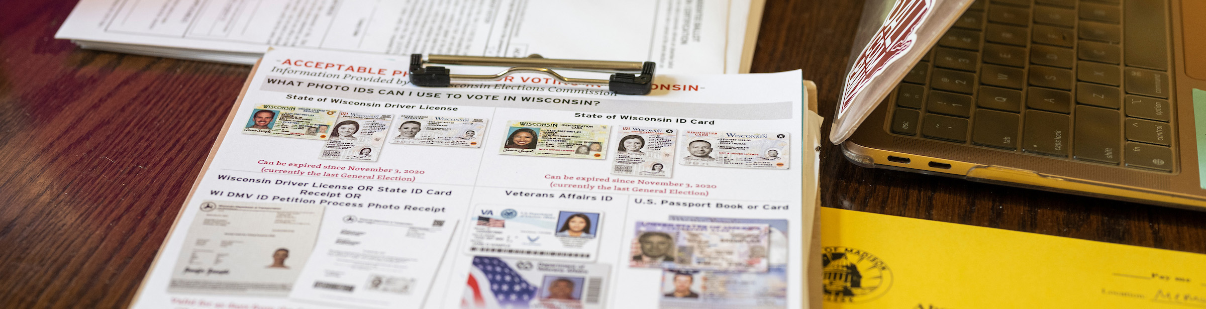 Photo of a clipboard sitting on a crowded table filled with forms. On the clipboard is an information sheet showing acceptable forms of voter identification.