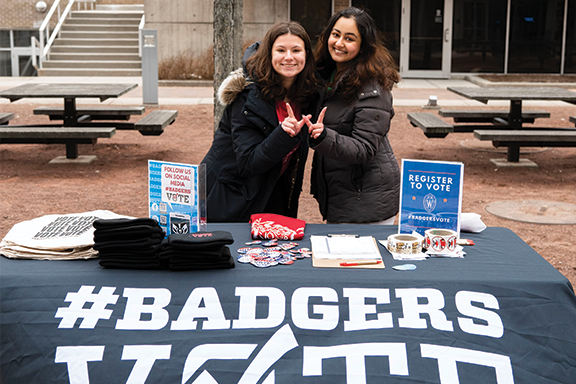 Two students stand behind a table with a black tablecloth with the words Badgers Vote printed on the front. They are holding their right hands together to make the Wisconsin W sign with their thumbs and index fingers.